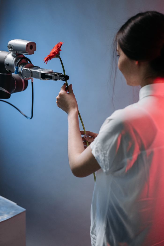 services-02 A robotic arm delicately holds a red flower as a woman interacts with it.
